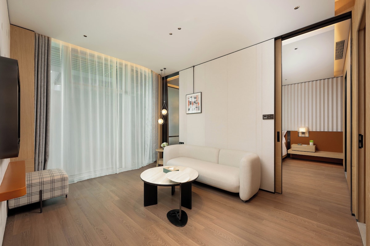 A modern hotel room with a sofa, a round coffee table, a TV, and a large window with curtains.