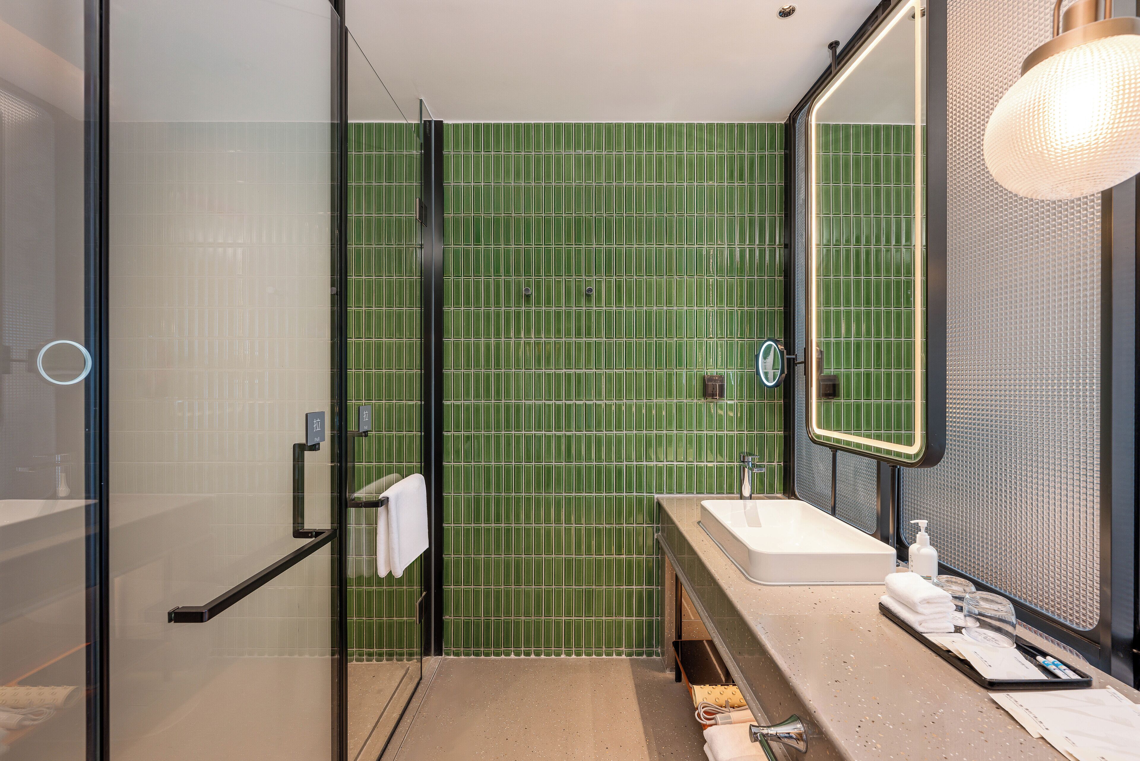 A modern bathroom with a glass shower enclosure, a sink with a mirror, and a green tiled wall.