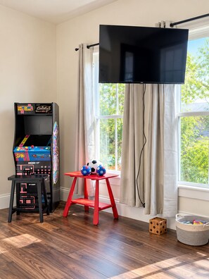 Une pièce avec un plancher en bois, un tabouret rouge orné de ballons de football, une borne d’arcade noire, un téléviseur fixé au mur et une fenêtre avec des rideaux.