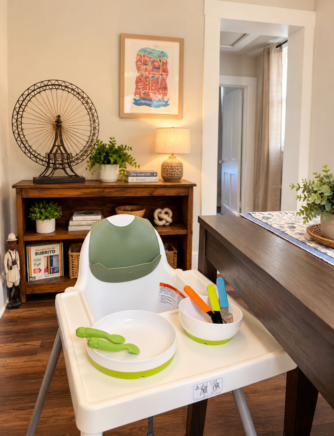 A dining area with a high chair, a bowl with utensils, a decorative lamp, a small plant, and a framed picture on the wall.