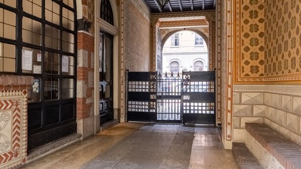Un pasillo con paredes de azulejos ornamentados, un portón negro y vista a un edificio a través de un arco.