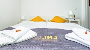 Ein Hotelzimmer mit einem Bett, ausgestattet mit einer gesteppten, grauen Bettdecke und zwei Kissen, eines gelb und eines weiß. Es gibt zwei weiße Nachttische mit Lampen und ein gerahmtes Bild an der Wand.