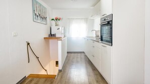 Eine schmale Küche mit weißen Schränken, einem eingebauten Ofen und einem Fenster mit durchsichtigen Vorhängen.