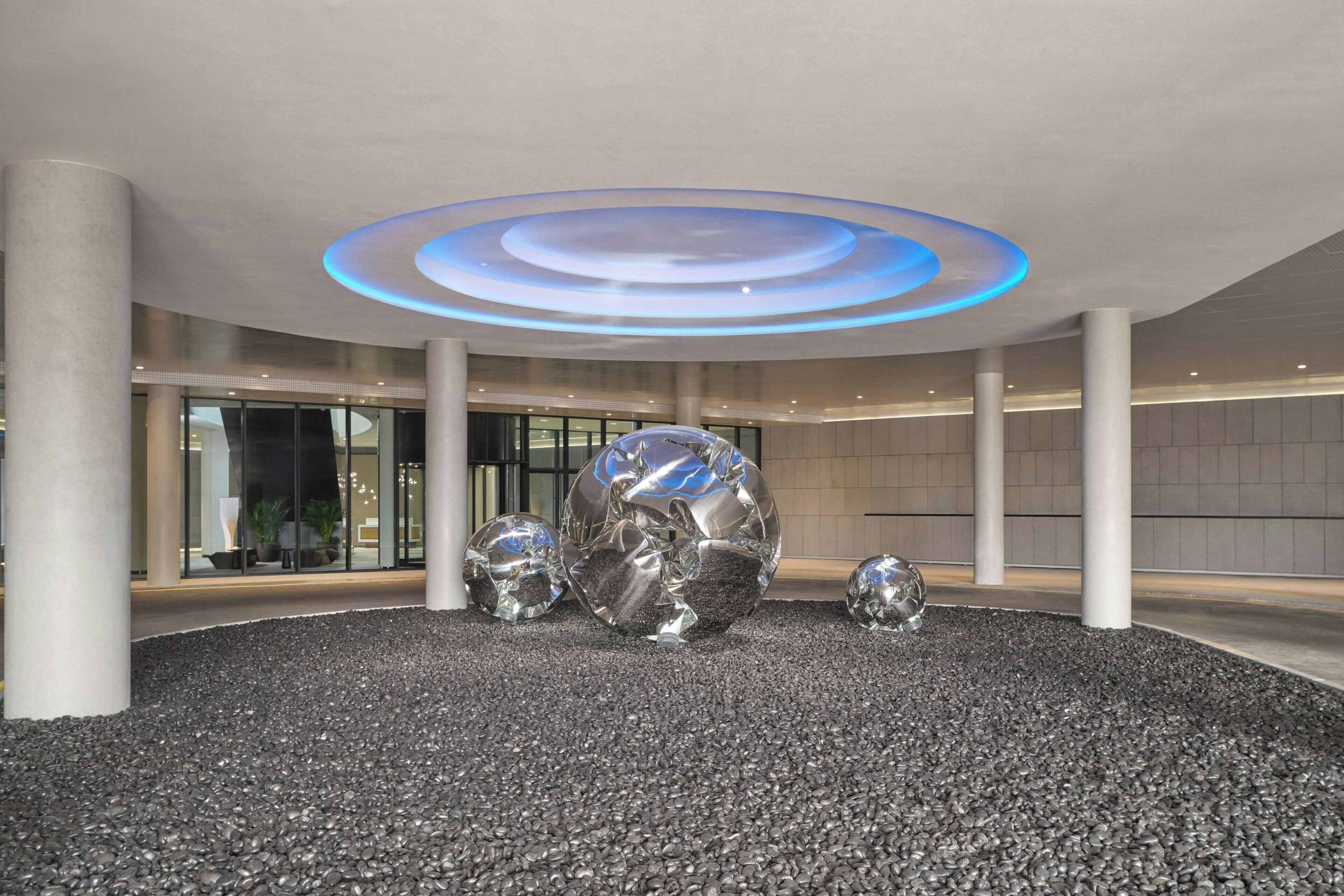 A modern lobby with a circular ceiling light, large metallic spheres, and a pebbled floor.