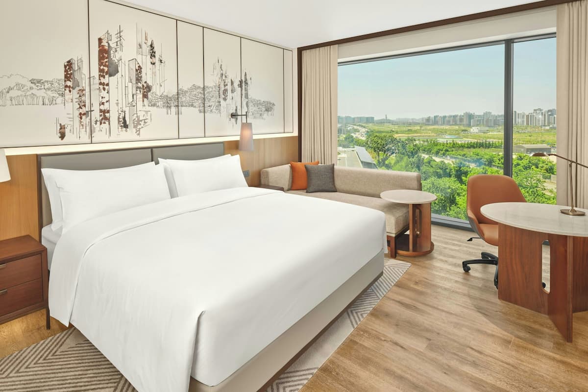 Habitación de hotel moderna con una cama grande, un sofá, una pequeña mesa redonda y vistas a un paisaje urbano y zonas verdes.