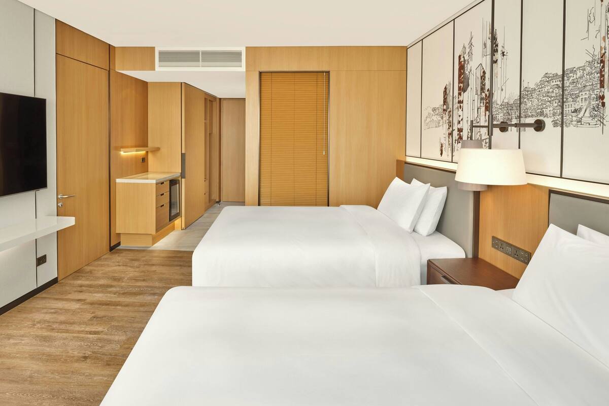 Habitación de hotel con dos camas, paneles de madera, un televisor de pantalla plana, una kitchenette pequeña y una obra de arte con vista a la ciudad.