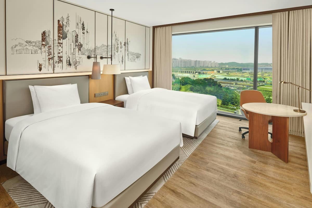 Habitación de hotel con dos camas, un escritorio y un amplio ventanal con vista a la ciudad.