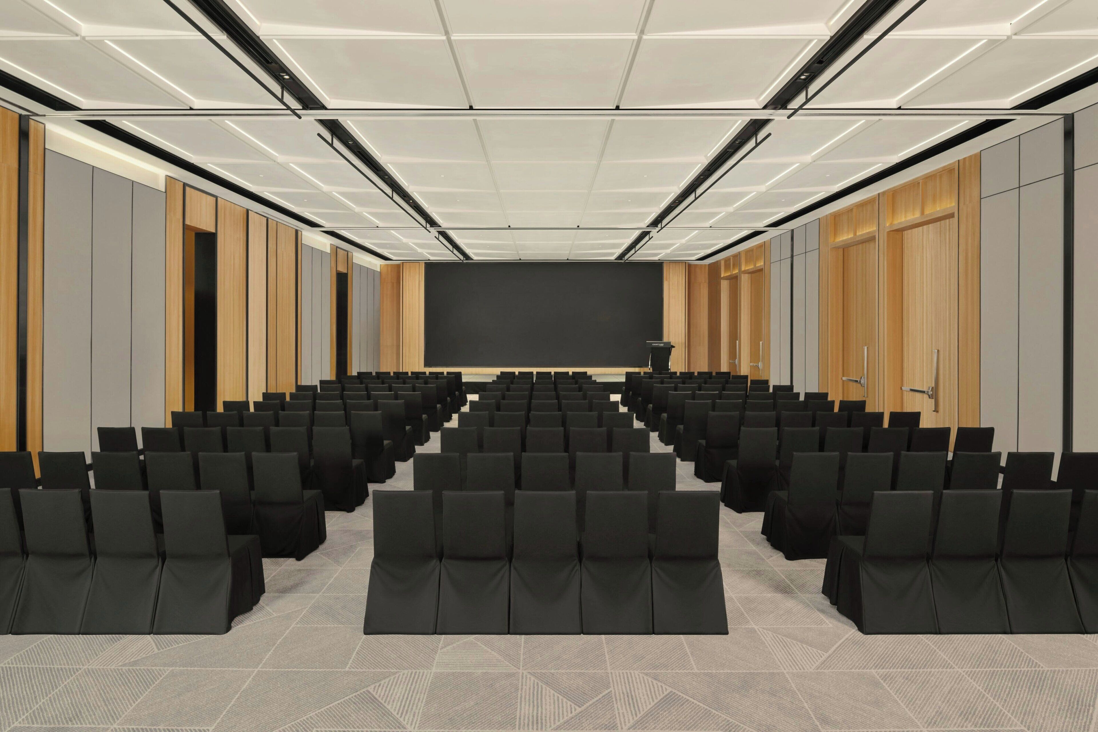Un salón de conferencias con filas de sillas, un escenario y un proyector.