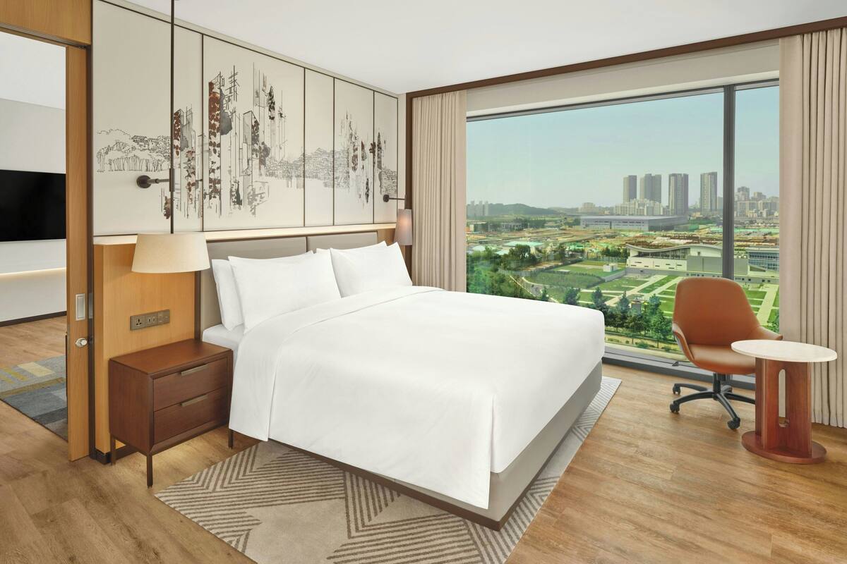 Una habitación de hotel moderna con una cama grande, una mesita de noche, un escritorio, una silla y un amplio ventanal con vistas a la ciudad.