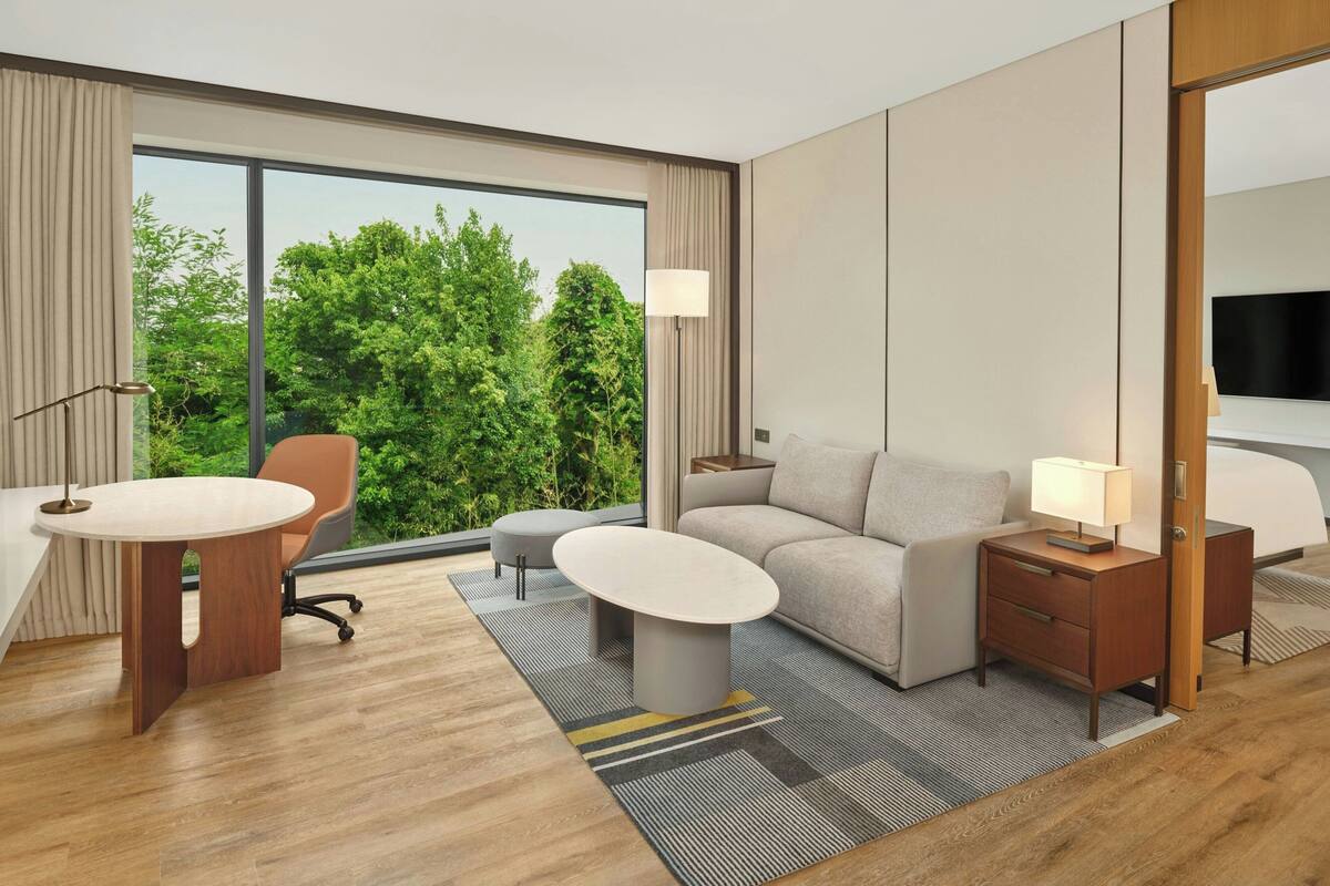 Una habitación de hotel moderna con un ventanal grande, un sofá, una mesa de centro, un escritorio y una mesita de noche.