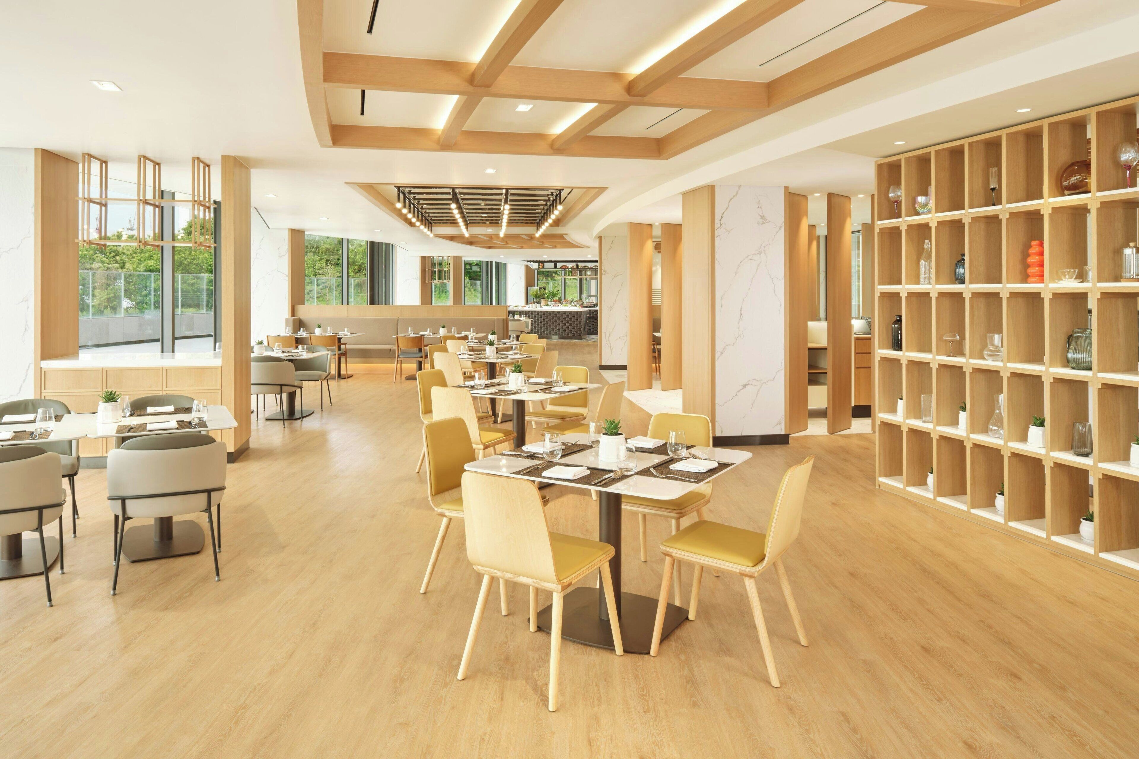 A spacious dining area with wooden tables and chairs, a large bookshelf, and a view of the outdoors through large windows.