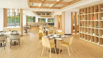 A spacious dining area with wooden tables and chairs, a large bookshelf, and a view of the outdoors through large windows.