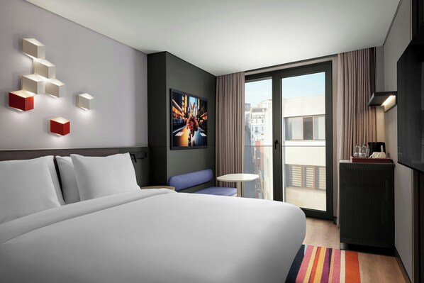 Habitación de hotel moderna con una cama grande, una zona de estar, una mesita, un cuadro y vistas a la ciudad a través de la ventana.