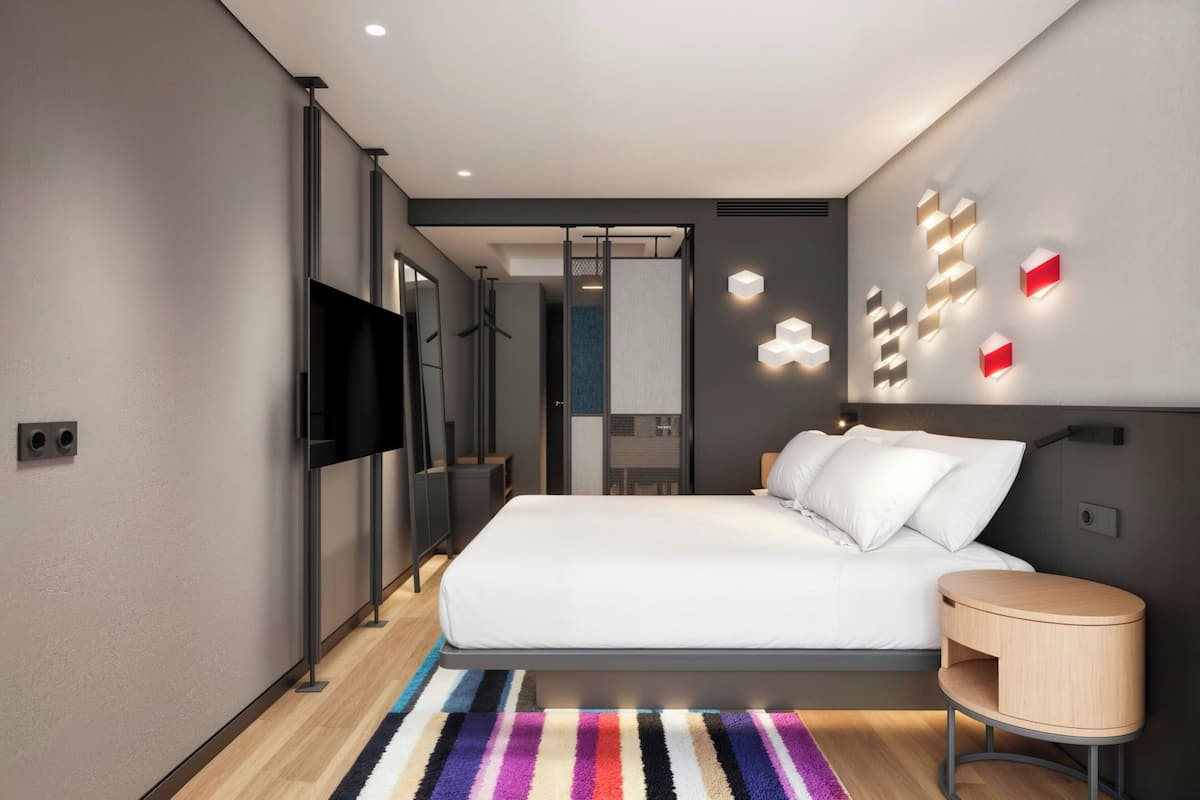 Una habitación de hotel moderna con una cama grande, una mesita de noche, un televisor y una lámpara de pared original.