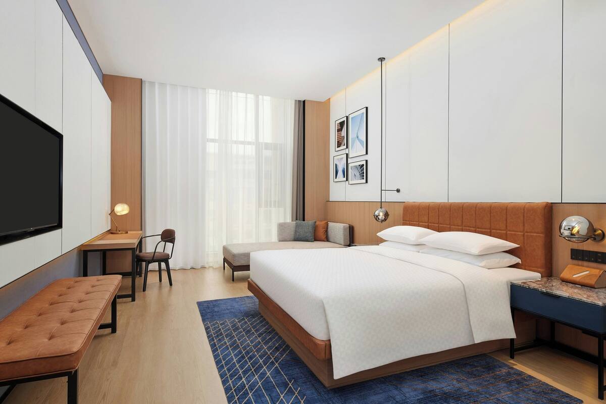 Ein modernes Hotelzimmer mit einem großen Bett, einem Schreibtisch, einem Stuhl, einer Bank, einem Fernseher und gerahmten Bildern an der Wand.