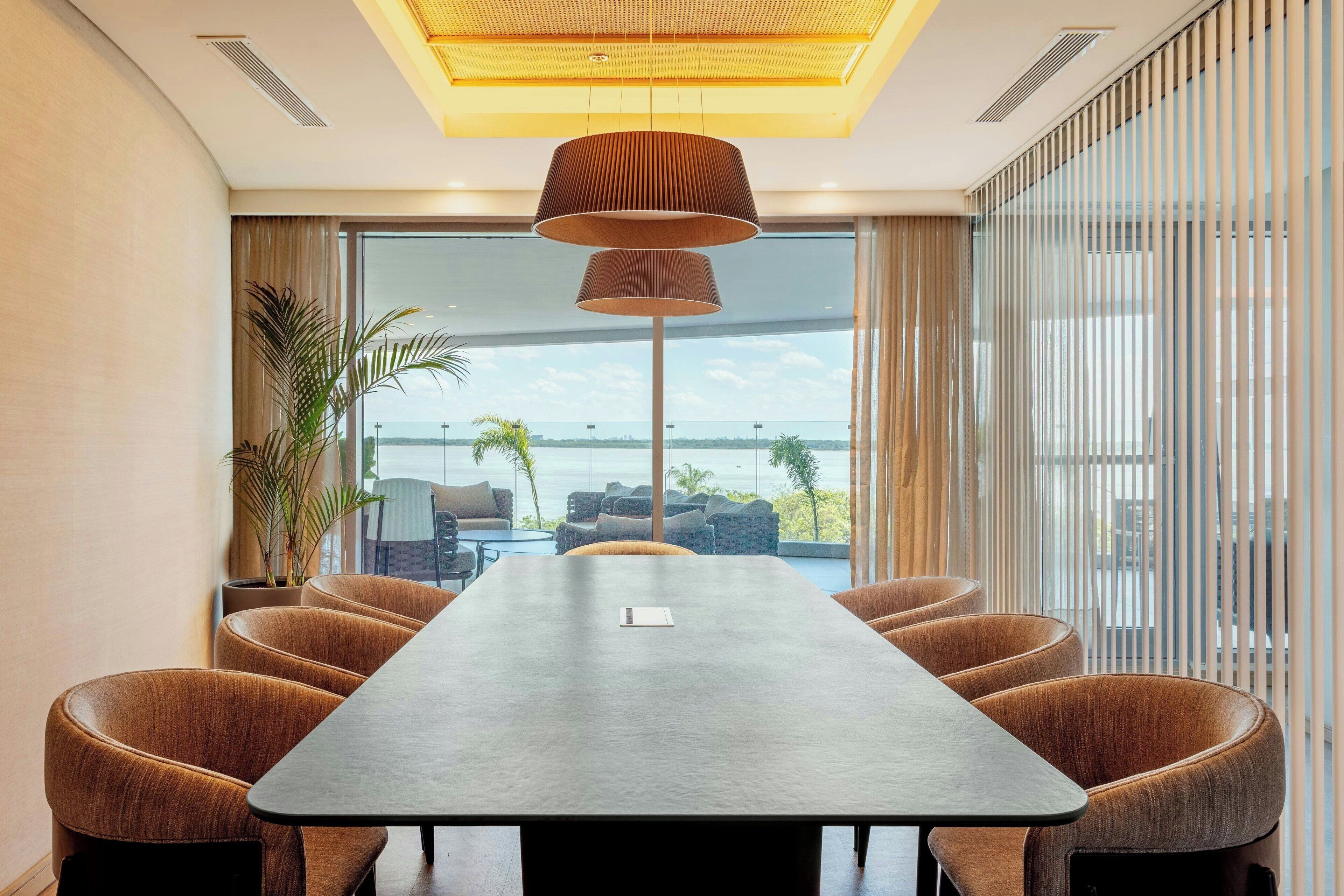 Un salón de conferencias con una larga mesa, sillas tapizadas en tonos marrones, una planta en maceta y vista al mar a través de amplias ventanas.
