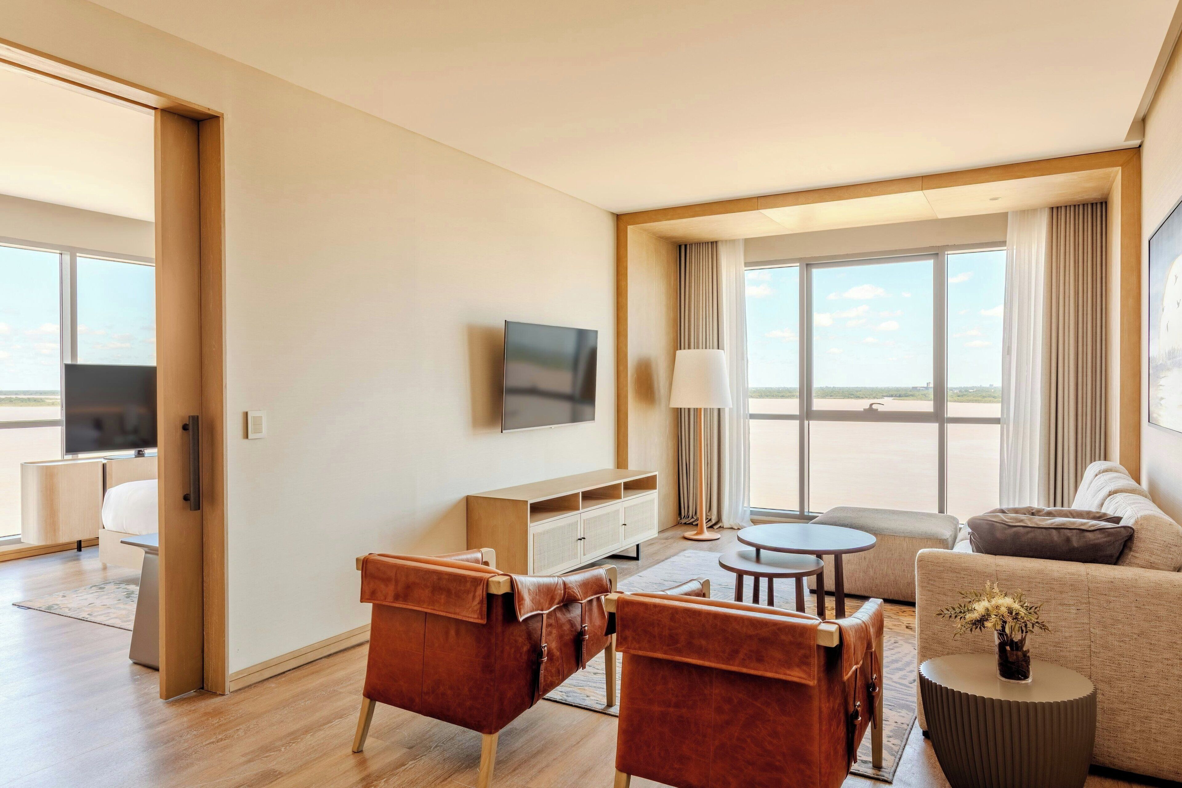 Ein modernes Hotelzimmer mit Fernseher, Ledersessel, einer Couch, einem kleinen runden Tisch und einem großen Fenster mit Blick ins Freie.