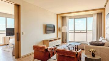 Ein modernes Hotelzimmer mit Fernseher, Ledersessel, einer Couch, einem kleinen runden Tisch und einem großen Fenster mit Blick ins Freie.