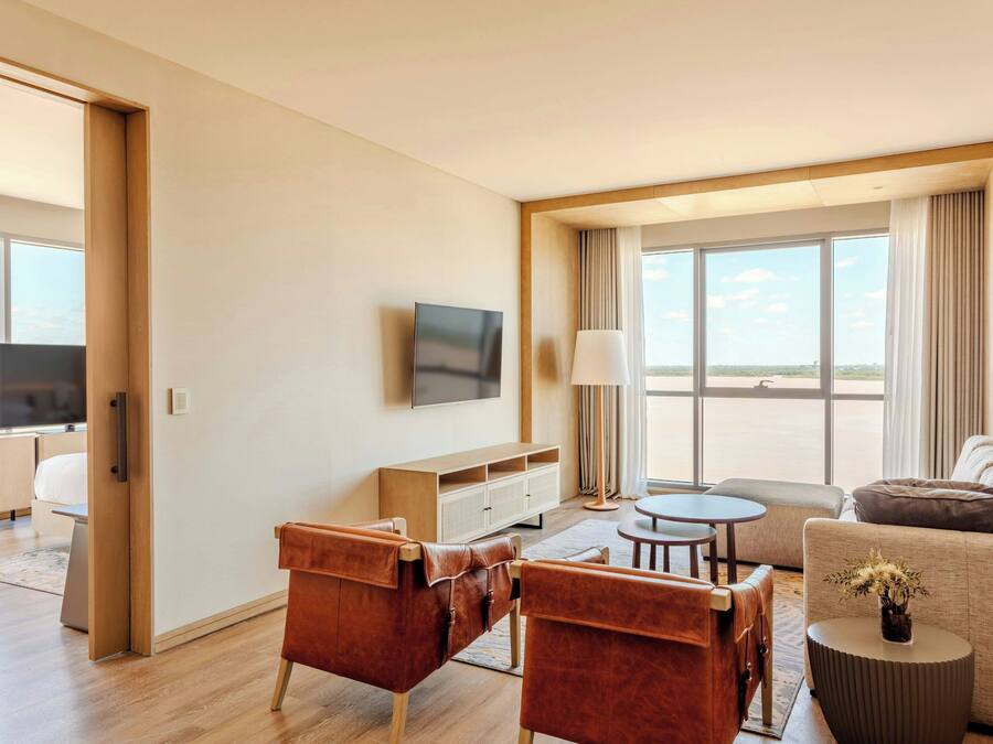 Ein modernes Hotelzimmer mit Fernseher, Ledersessel, einer Couch, einem kleinen runden Tisch und einem großen Fenster mit Blick ins Freie.