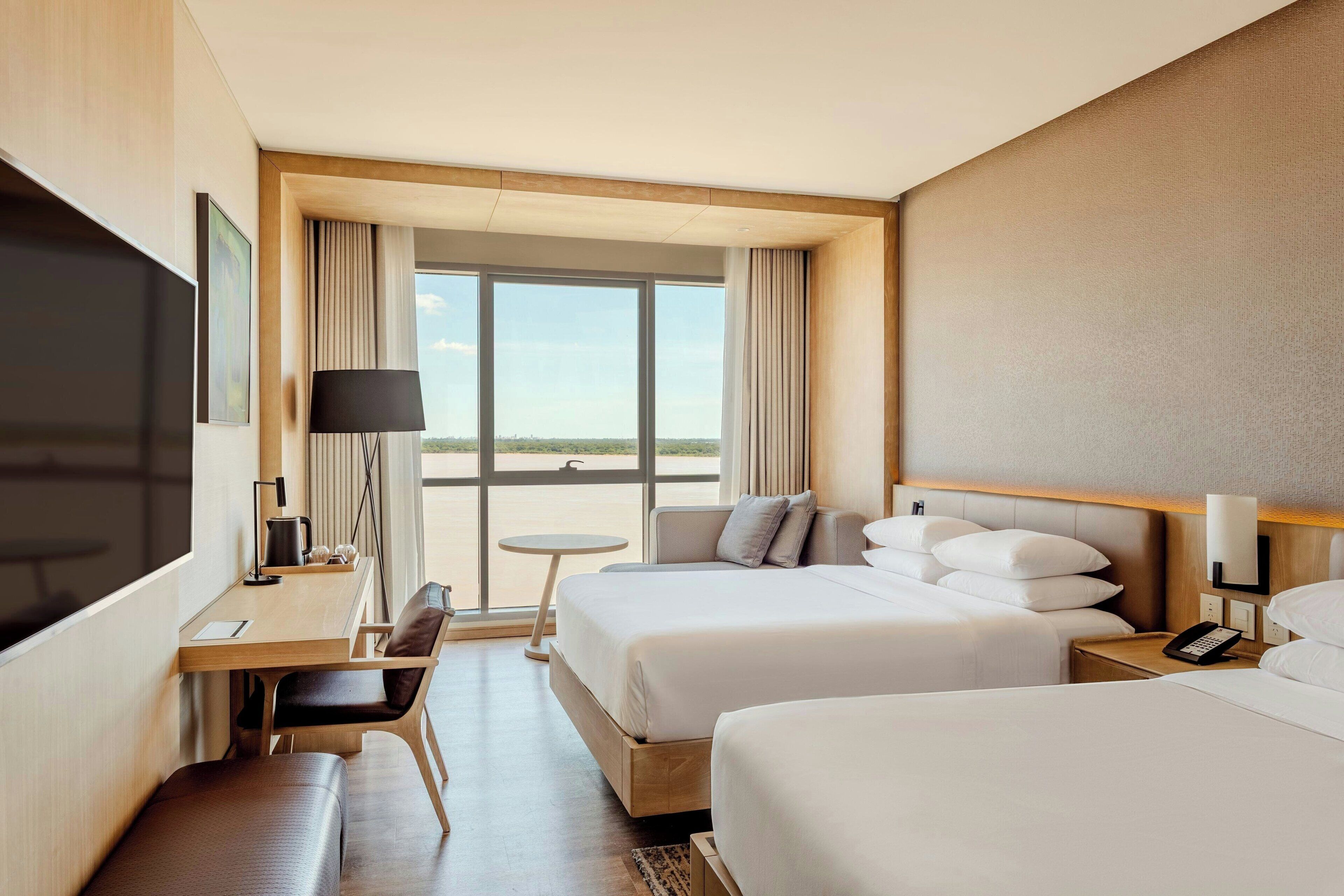 Habitación de hotel con una cama grande, una cama más pequeña, un escritorio con silla, un televisor y vistas al exterior.