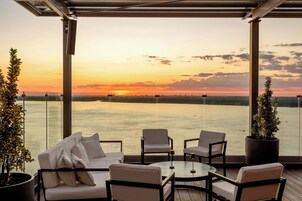 Una terraza con vistas a un atardecer sobre el agua, equipada con un sofá, sillas y una mesita.