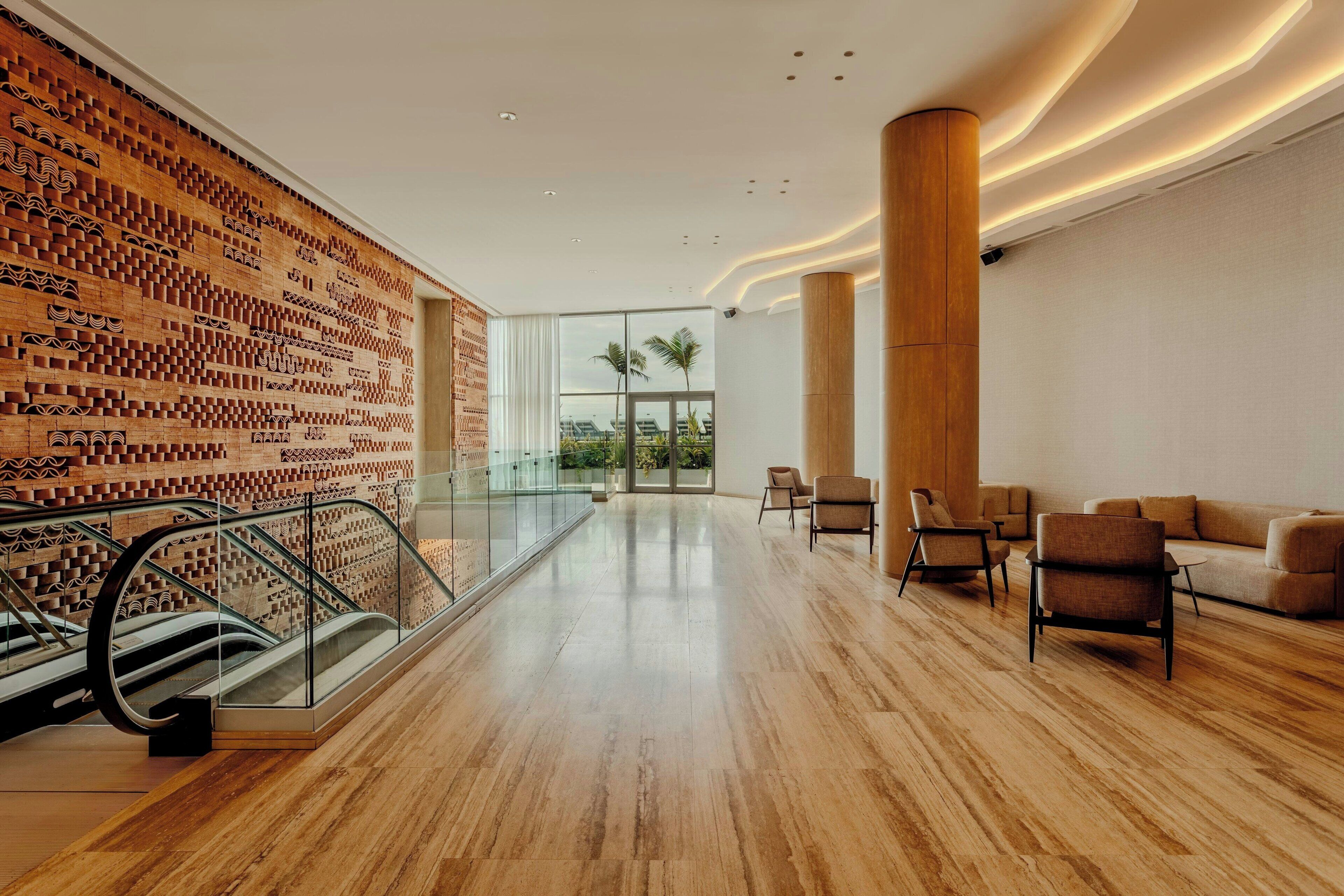 Un vestíbulo amplio con piso de madera, pared decorada, baranda de vidrio y zona de estar con sillas y un sofá.