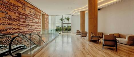 Un vestíbulo amplio con piso de madera, pared decorada, baranda de vidrio y zona de estar con sillas y un sofá.