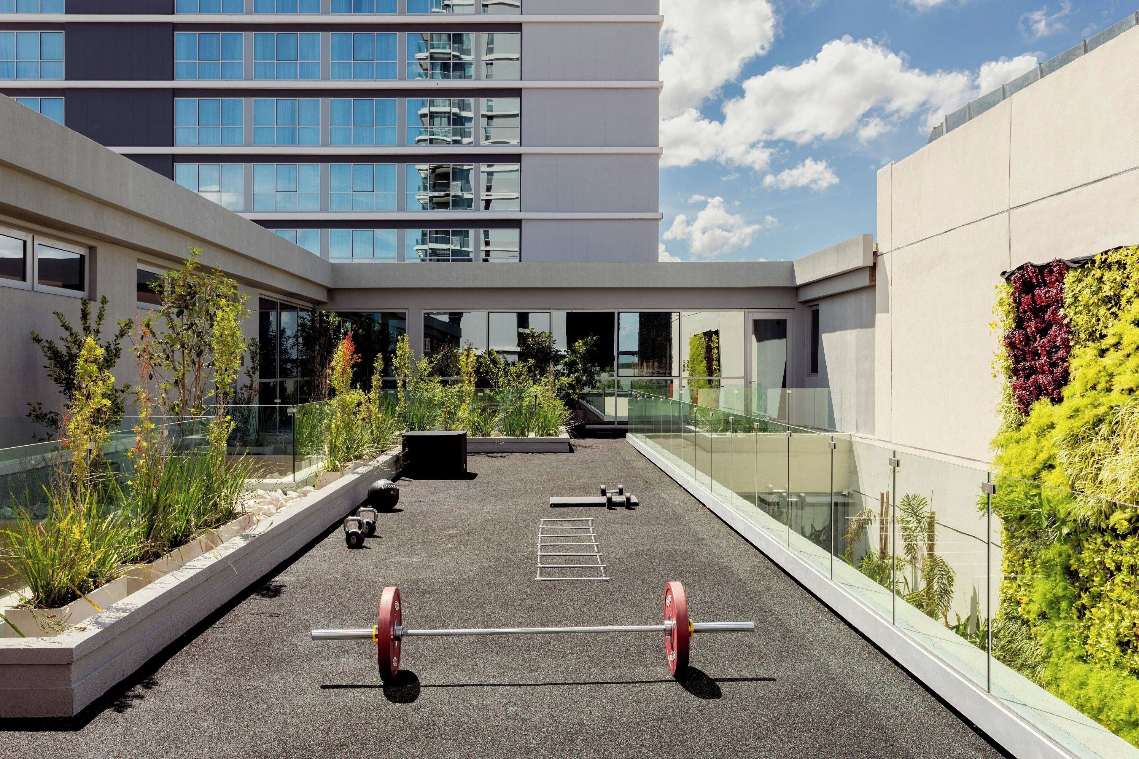 A rooftop gym with outdoor exercise equipment, surrounded by greenery and modern buildings.