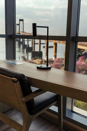 A wooden desk with a black chair, a tablet, and a black stand, positioned by a large window overlooking a body of water and buildings.