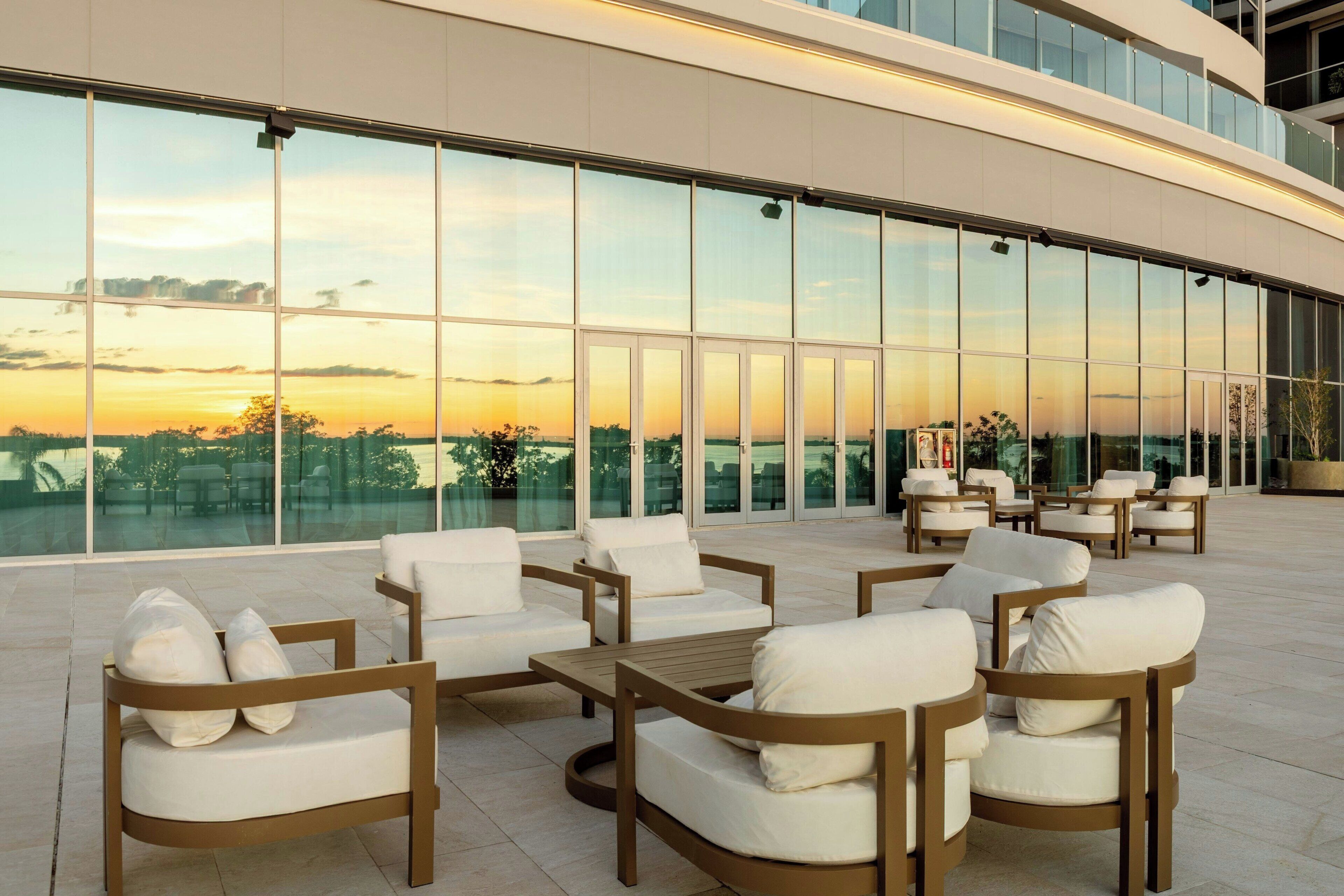 Un espacio exterior moderno con varios asientos y mesas, ventanas de vidrio amplias y vistas al paisaje.