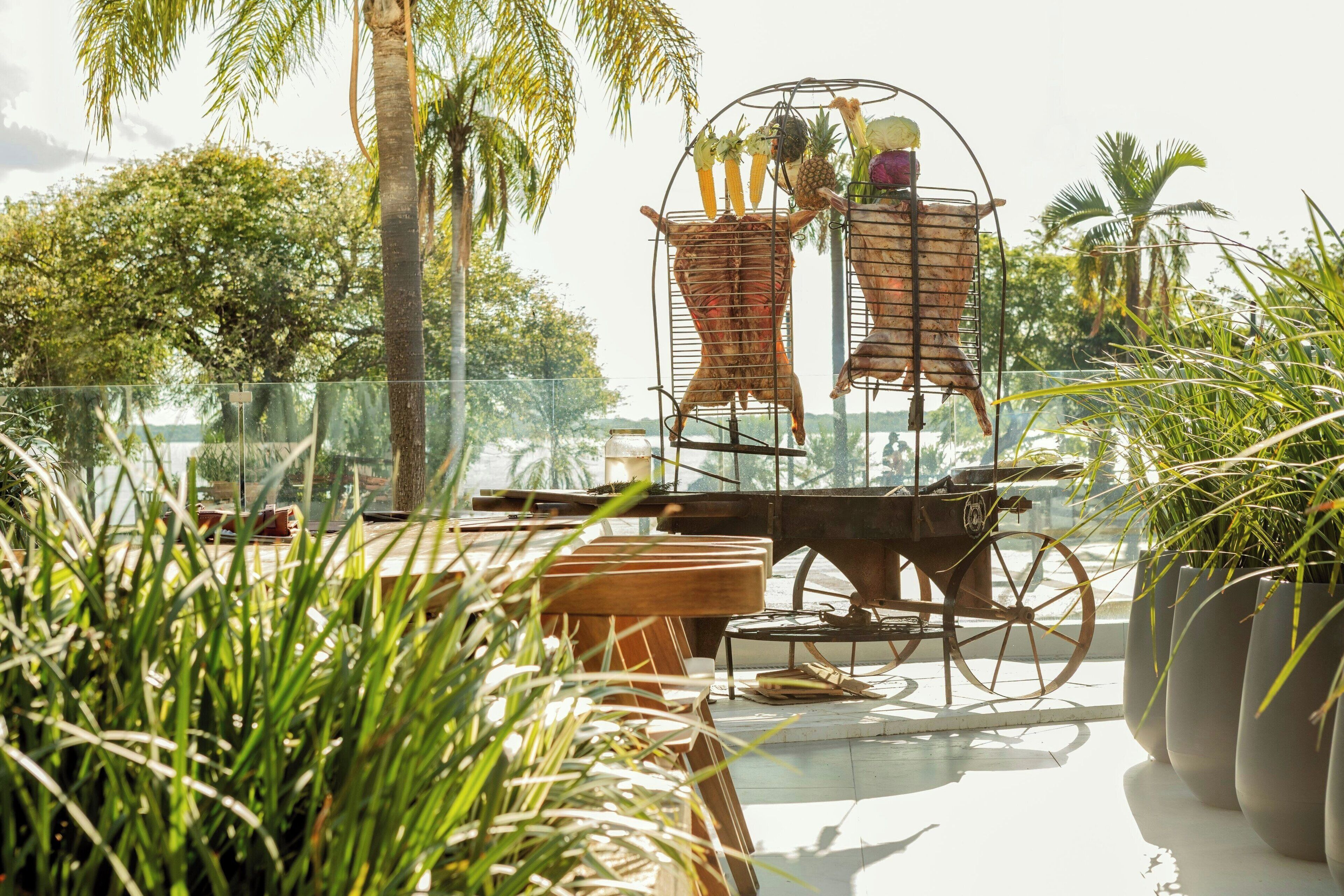 Un espacio al aire libre tropical con cerramiento de vidrio, un carrito de madera y diversas plantas.