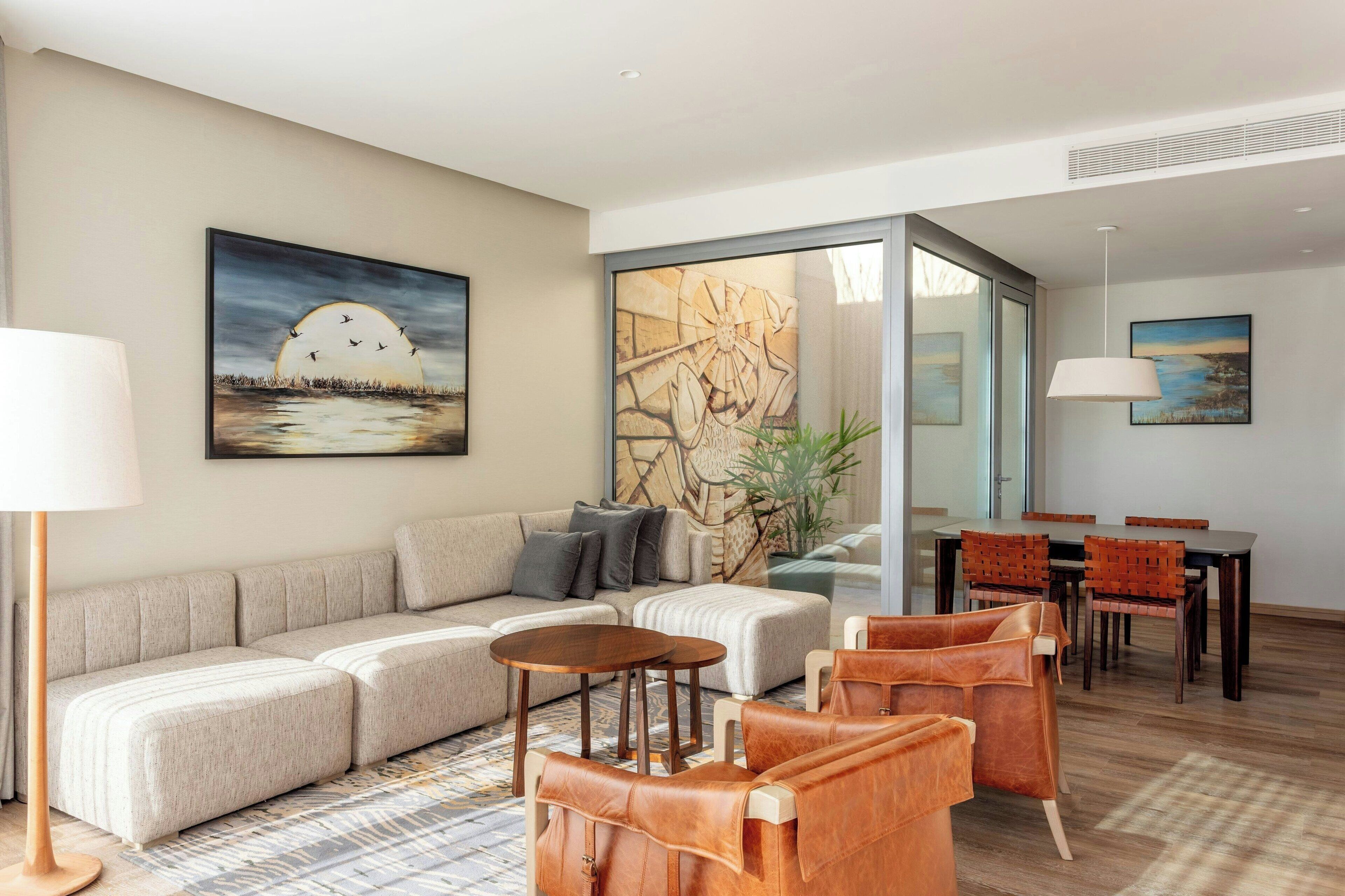 A modern living room with a sofa, coffee table, and leather chairs, a dining area with a table and chairs, and artwork on the walls.