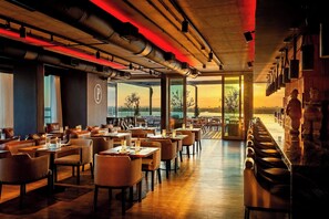 Un restaurante con una decoración moderna, que cuenta con asientos de cuero, una zona de bar con taburetes y grandes ventanales con vistas al exterior.