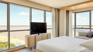 A hotel room with a large bed, a TV on a wooden stand, and a view of a beach and water through large windows. - Marriott Corrientes Hotel (Corrientes)