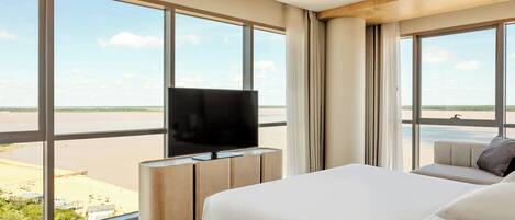 Une chambre d’hôtel avec un grand lit, un téléviseur posé sur un meuble en bois, et une vue sur la plage et la mer, visible à travers de grandes fenêtres.