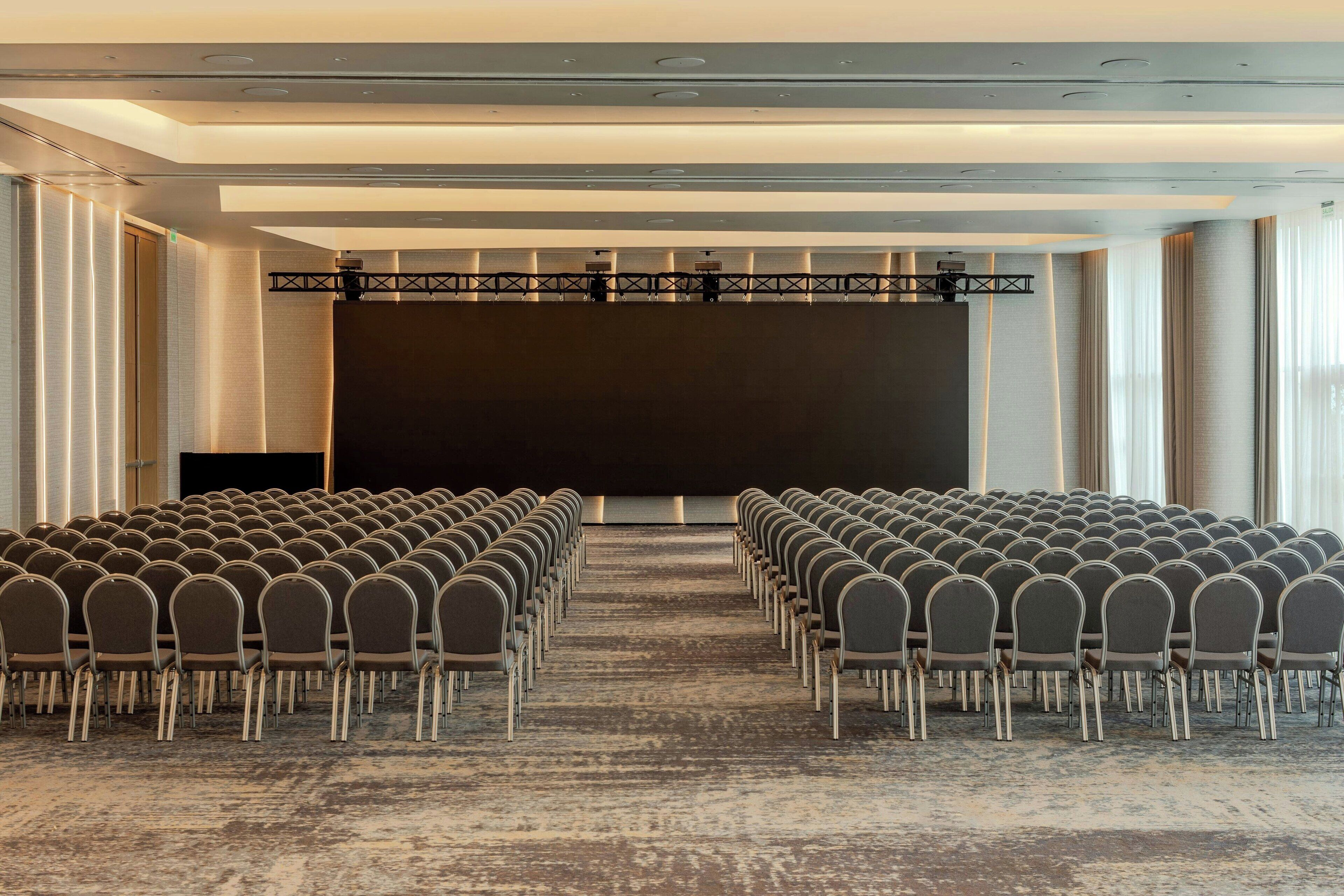 Ein Konferenzraum mit Reihen von Stühlen, die auf einen großen Bildschirm zeigen, mit Teppichboden und einer Bühne mit Beleuchtung.