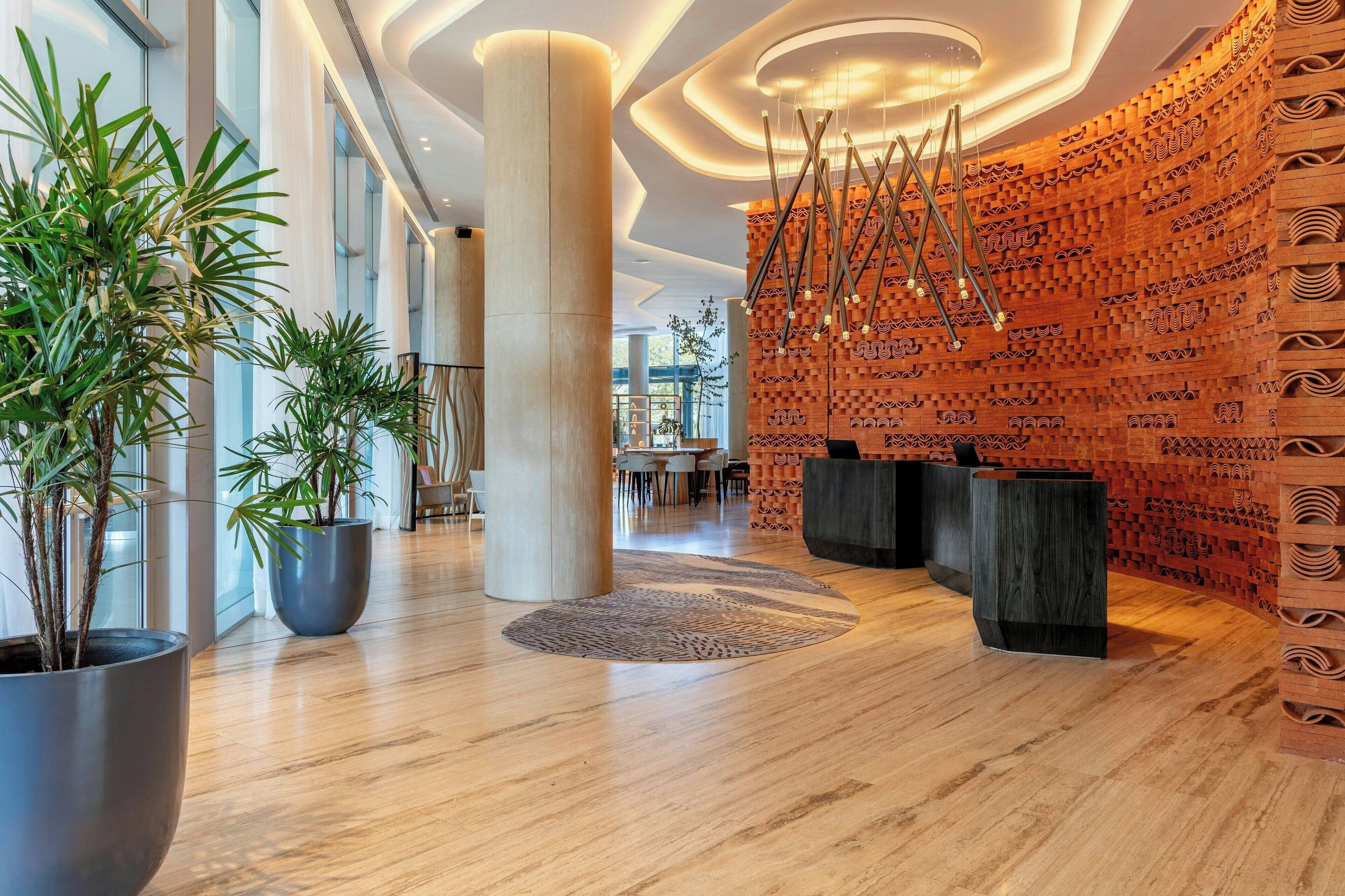 Un vestíbulo moderno con suelo de madera, grandes plantas en macetas, alfombra estampada y una pared con textura similar al ladrillo.