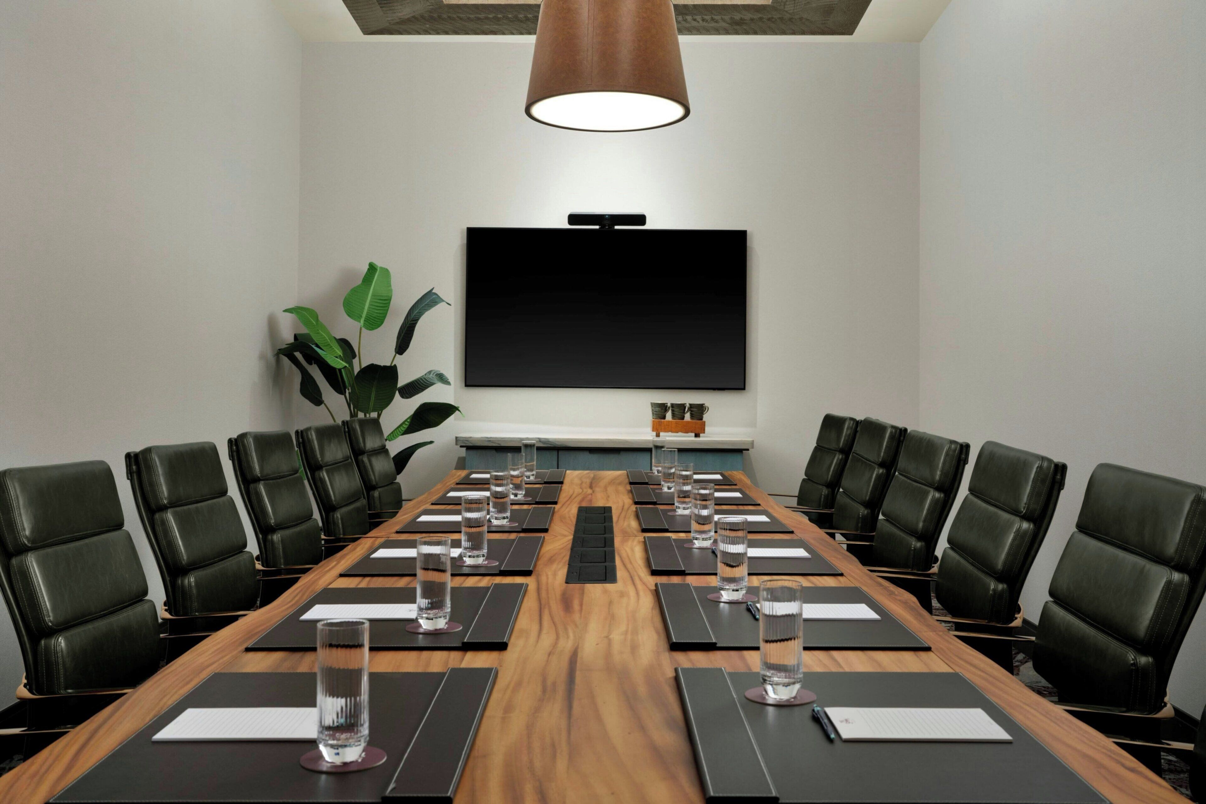 A conference room with a long table, black leather chairs, water glasses, and a flat-screen TV on the wall.