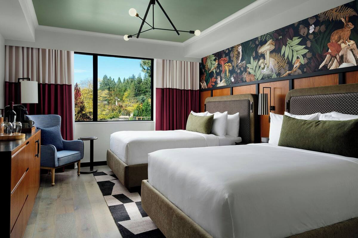 A hotel room with two beds, a desk, a chair, and a large window offering a view of trees.