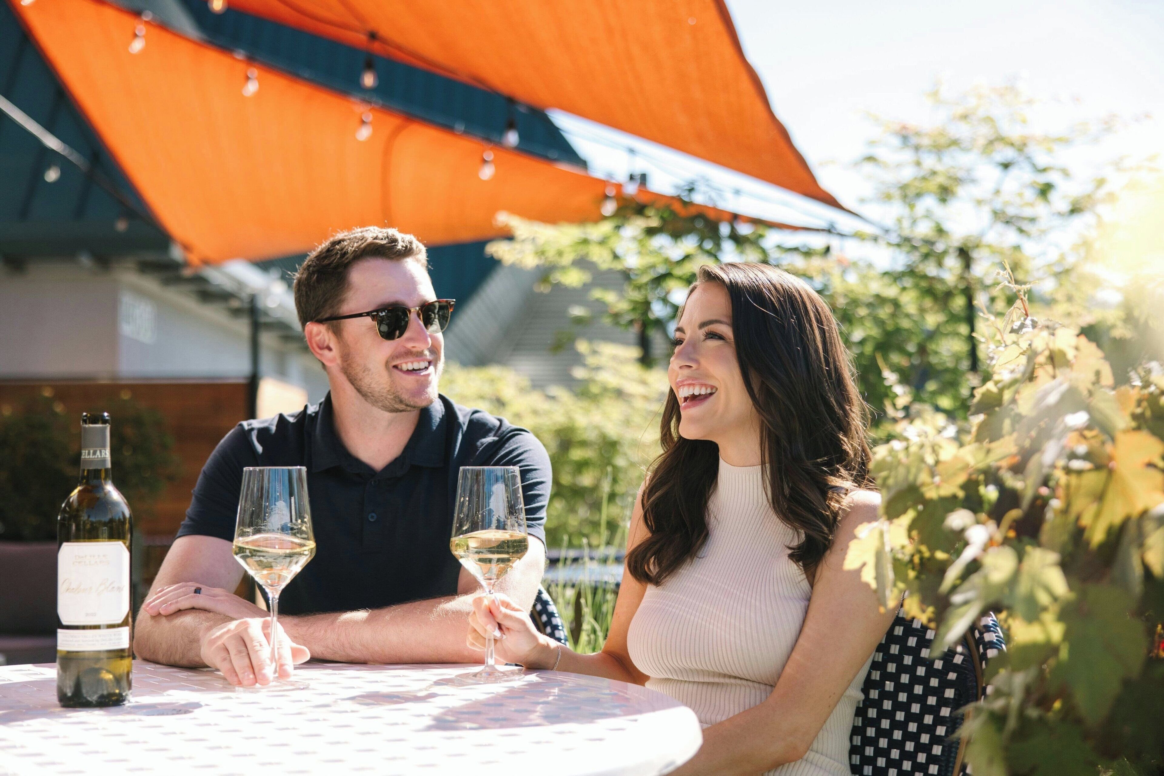 Un couple est assis à une table en plein air, devant une bouteille de vin et des verres.