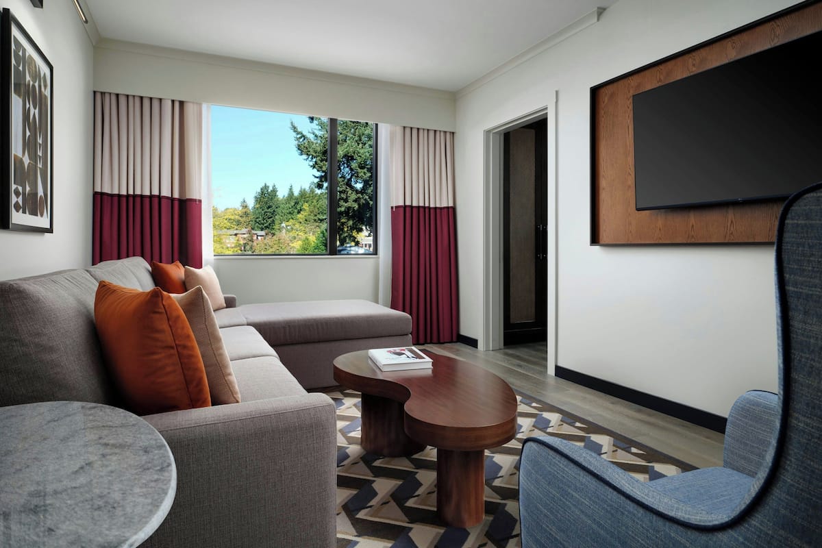 A modern living room with a sofa, armchair, and coffee table, a flat-screen TV mounted on the wall, and a large window with curtains.
