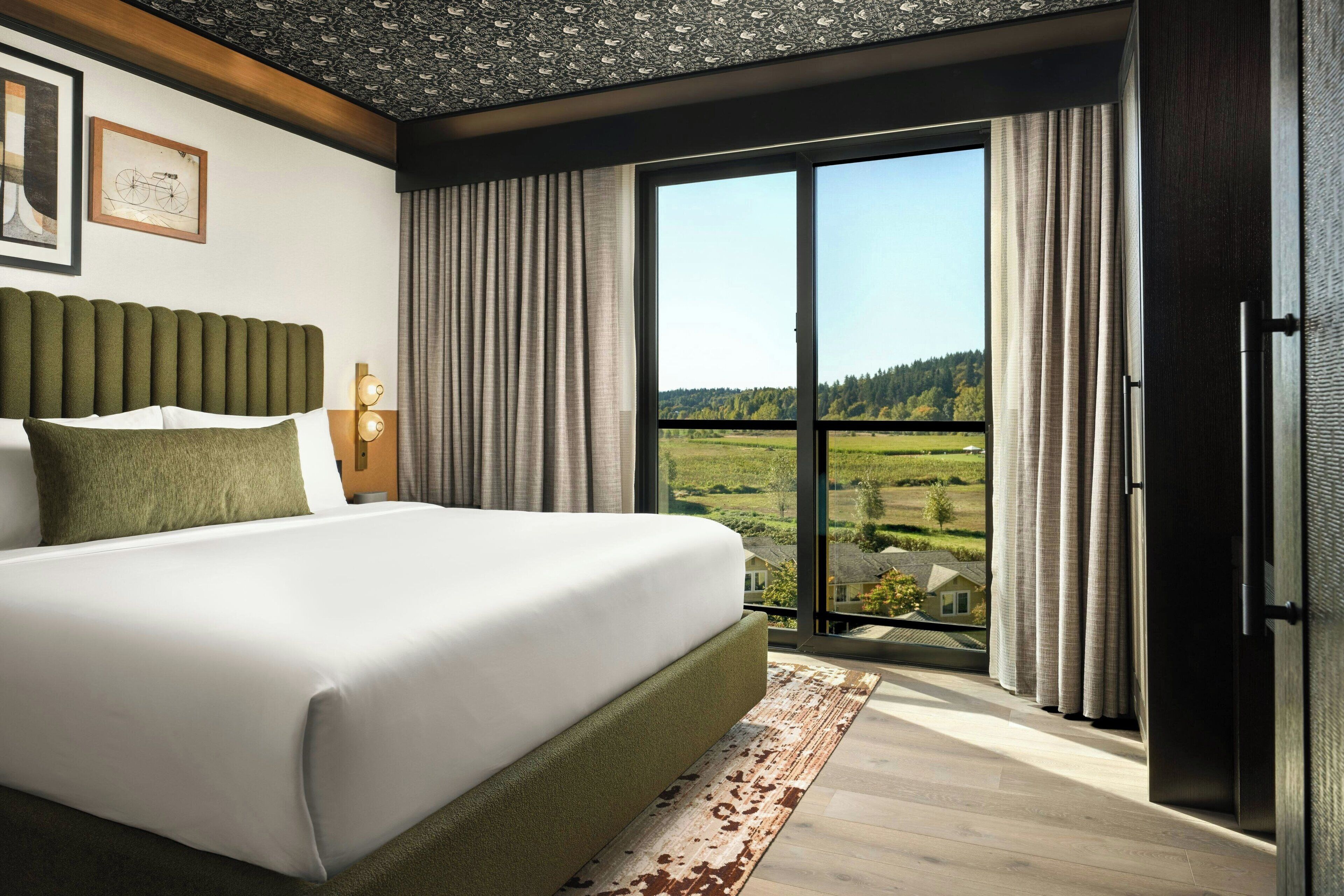 Una habitación de hotel moderna con una cama grande, vistas al campo a través de un ventanal corredera y techo de madera.
