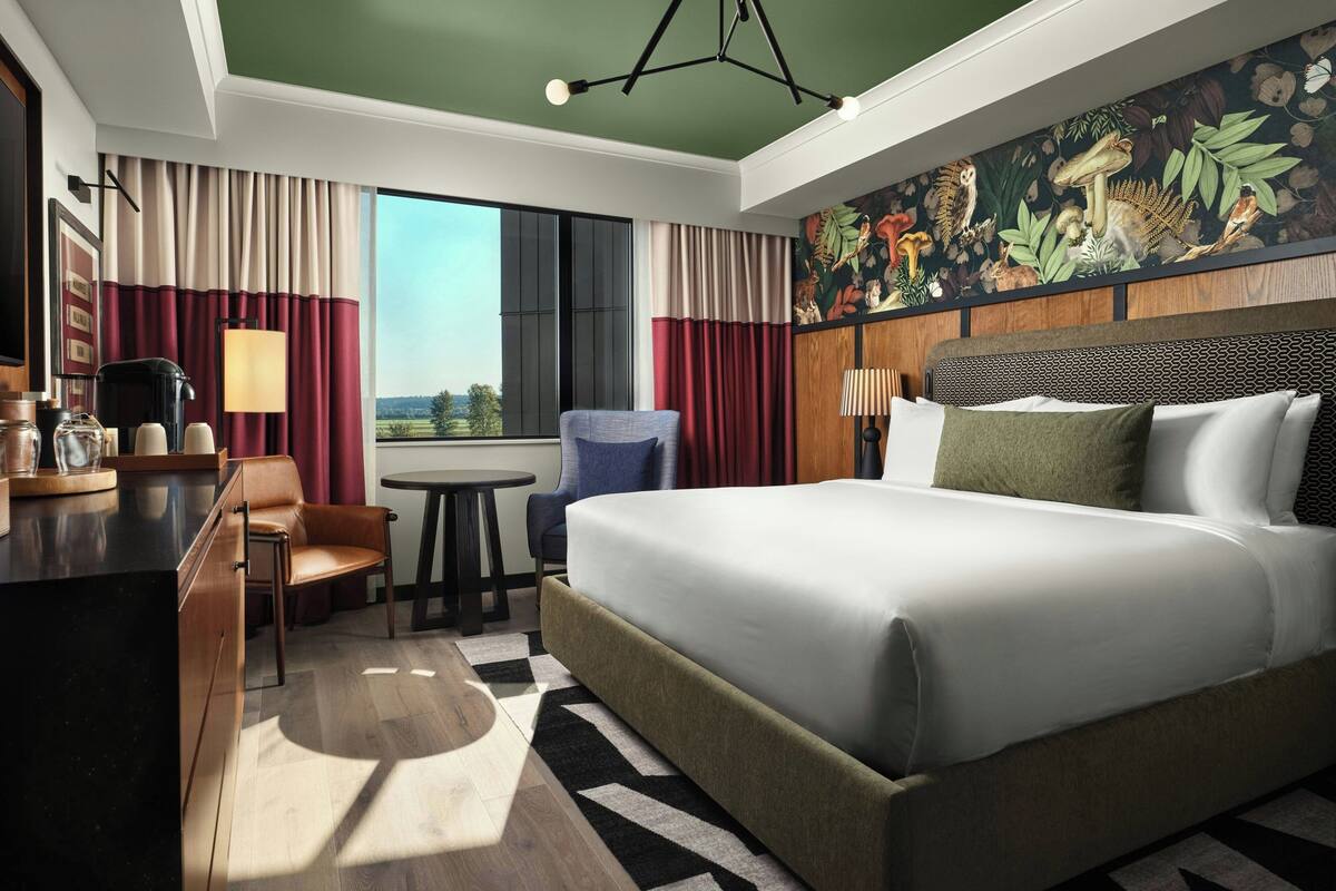 A hotel room with a large bed, a desk with a coffee maker, a chair, a small table, and a view of the outdoors.
