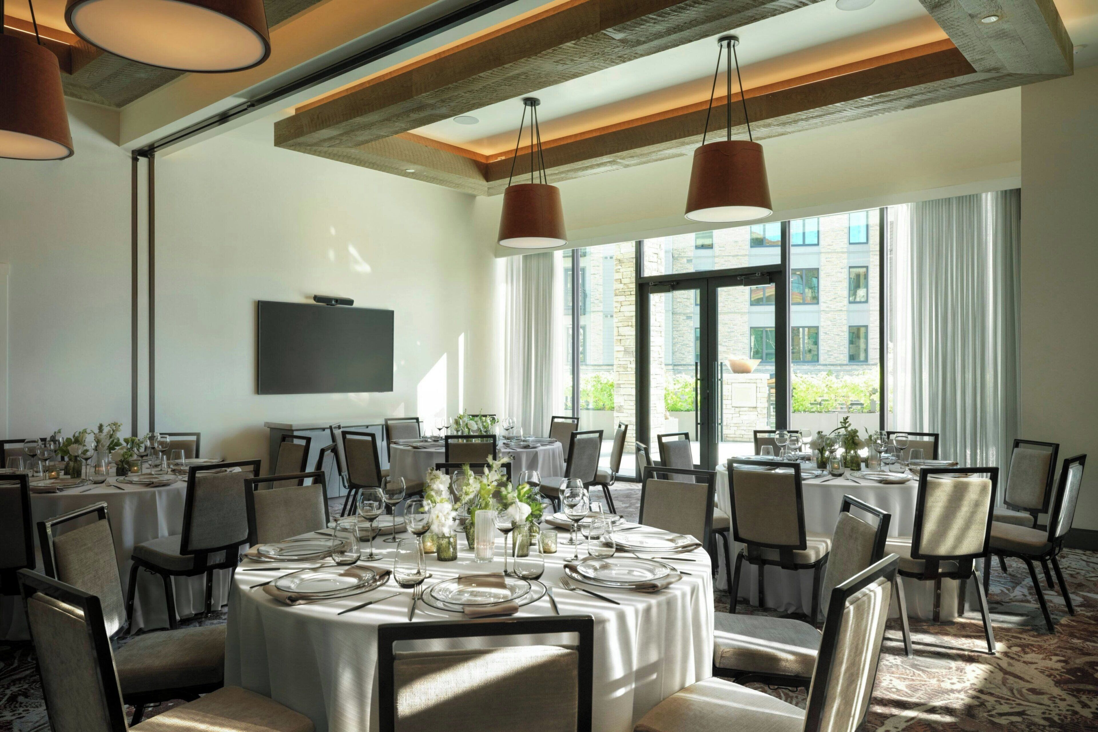 A well-lit dining room with round tables set for a meal, large windows with curtains, and a flat-screen TV mounted on the wall.