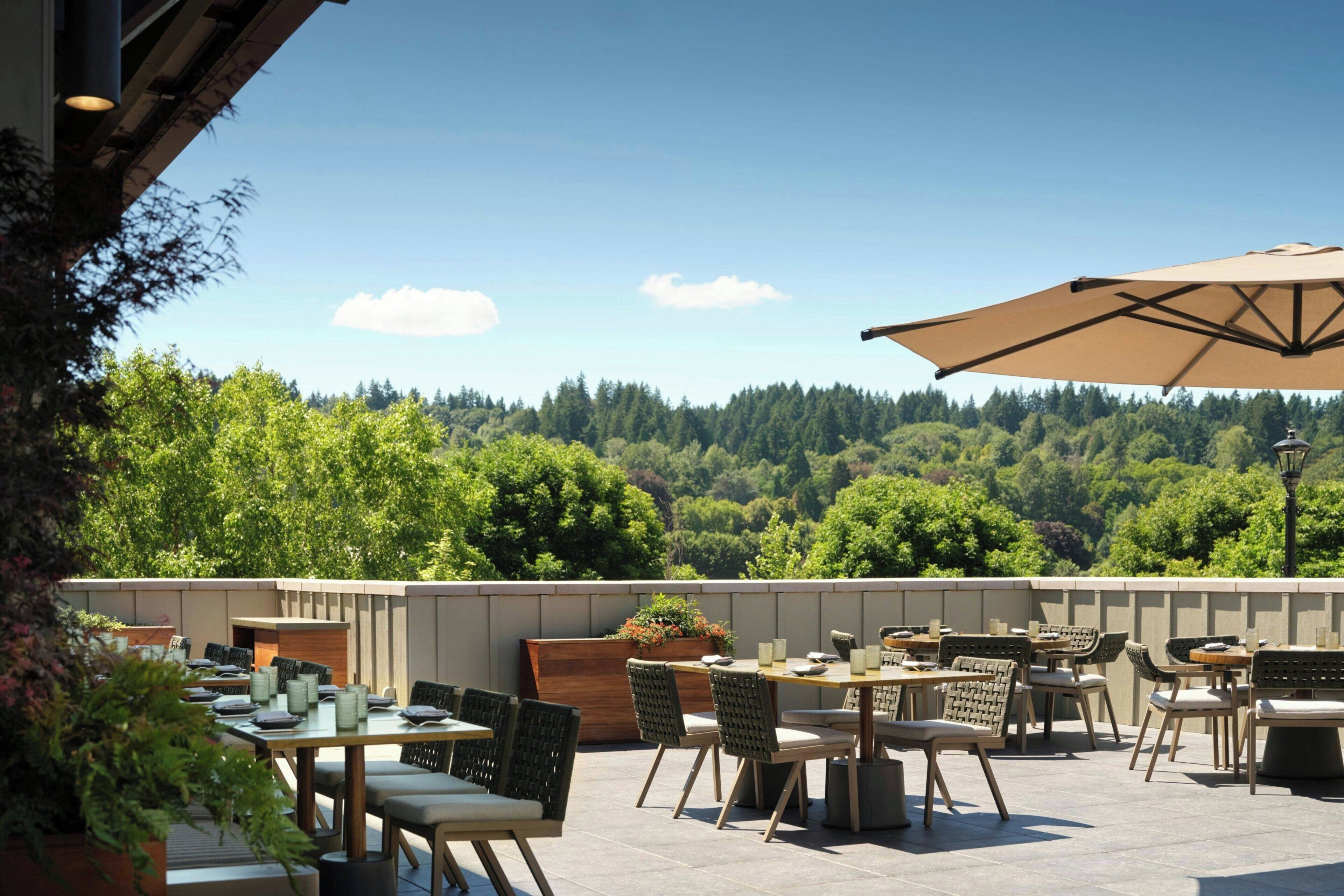 Une terrasse extérieure avec des tables et des chaises, un grand parasol et une vue sur une colline boisée.