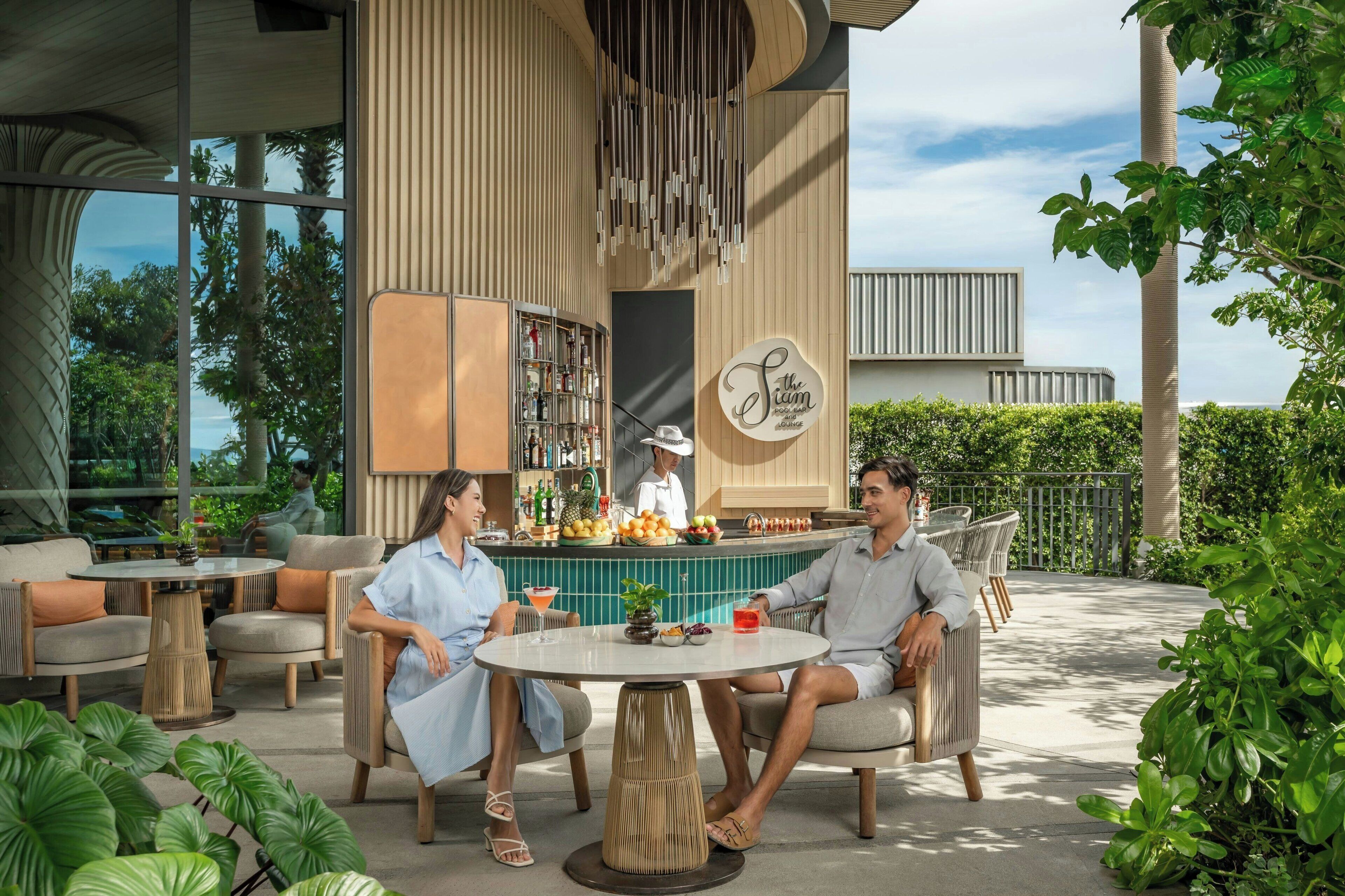 An outdoor seating area with round tables and chairs, a bar with a bartender, and a view of the surrounding greenery.