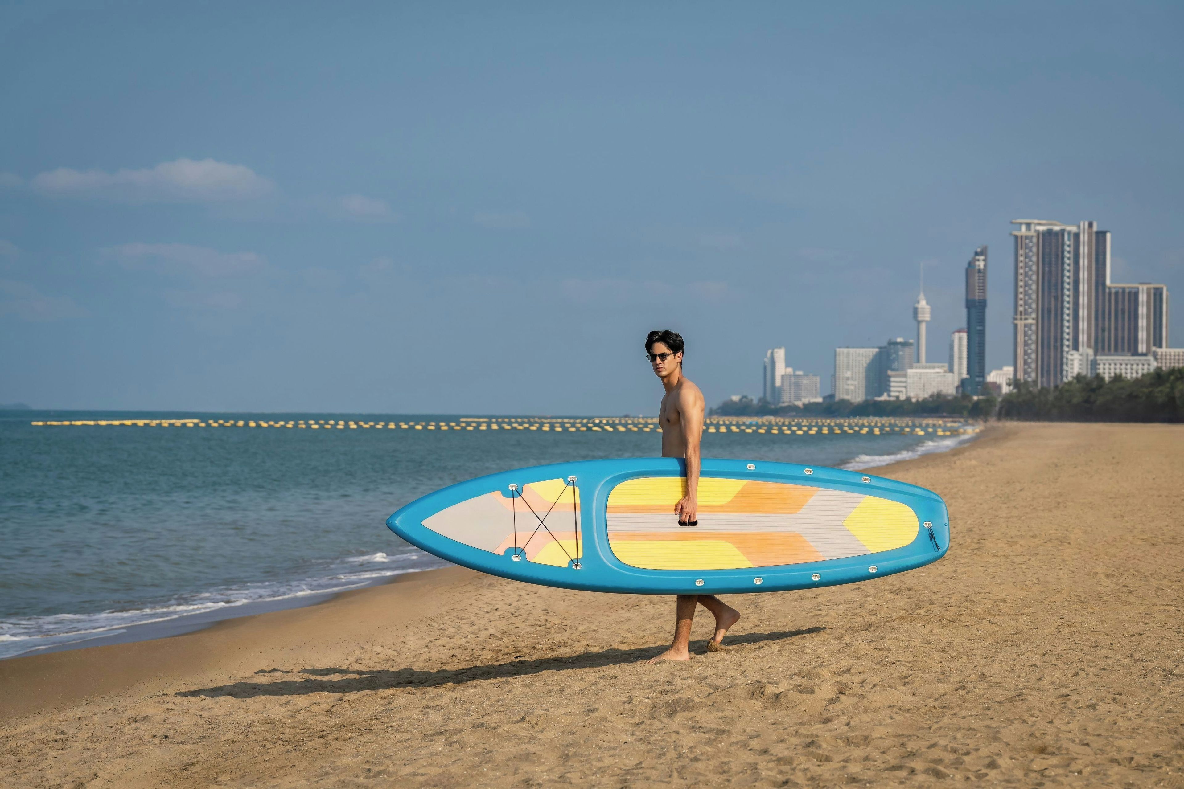 A person carrying a surfboard on a beach with a city skyline in the background.