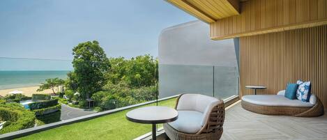 A balcony with a view of the beach, a wicker chair, a round table, and a cushioned seating area.