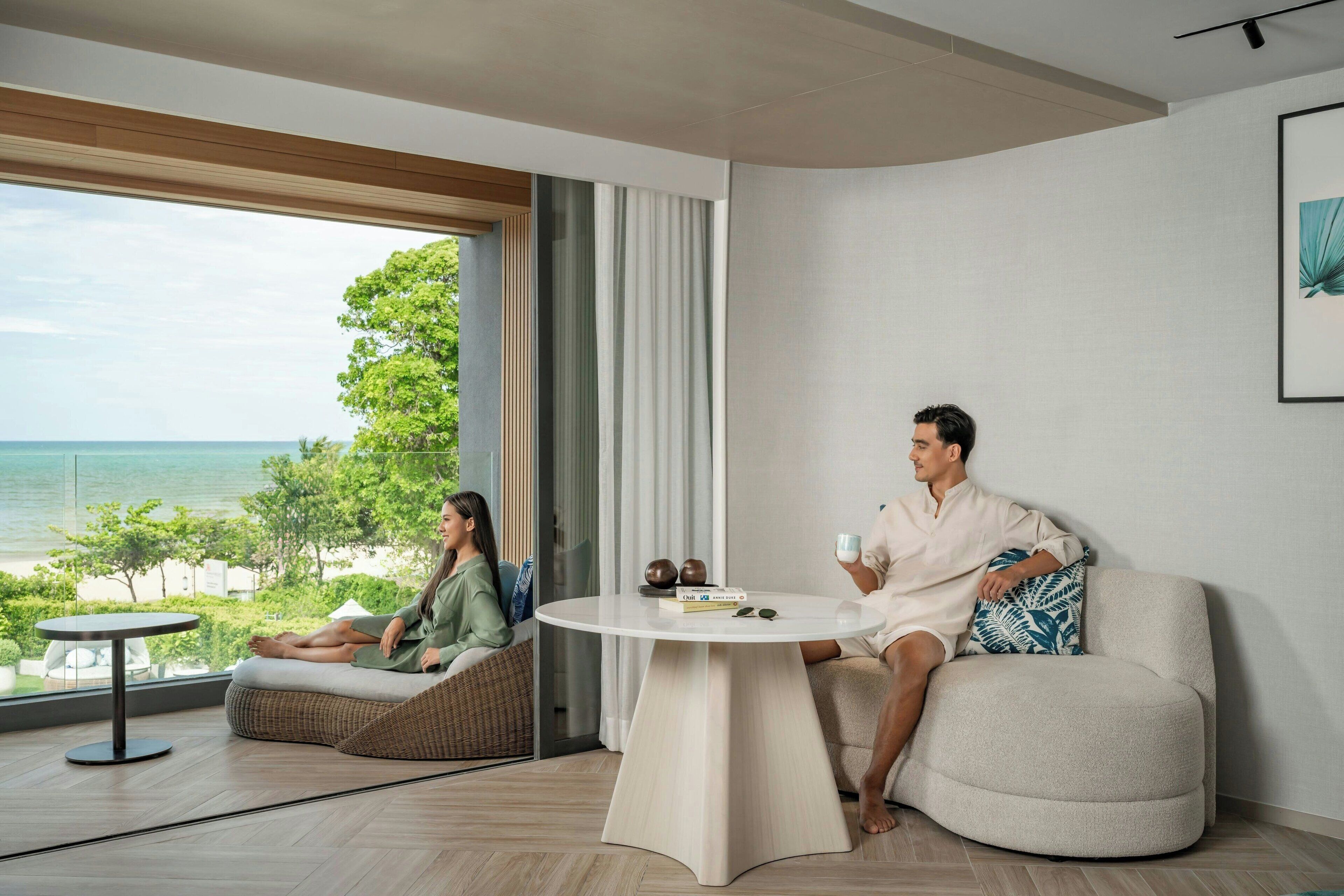 A modern living room with a large window overlooking a beach, a round coffee table, and a sofa with cushions.