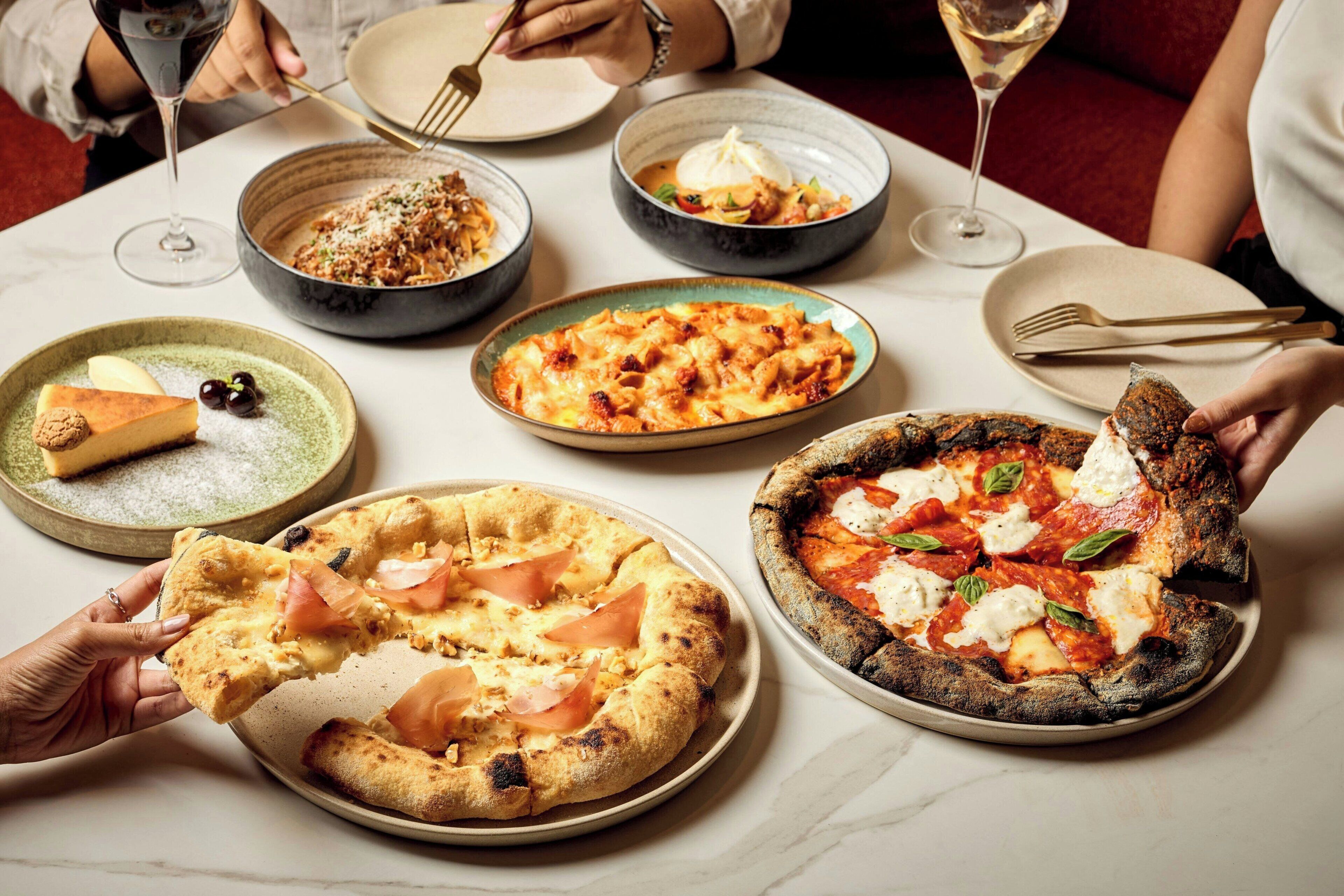 A dining table with various dishes, including pizza and pasta, and a person holding a slice of pizza.